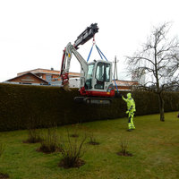 Mini-Bagger wird mit Greifarm über eine Hecke gehoben